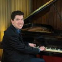 Jason Coleman in a black suit jacket, turned smiling at the camera, hands on the piano in front of him