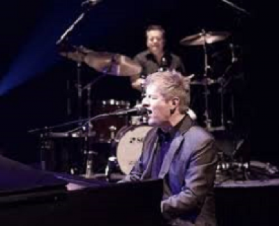 Jim sitting at a piano singing with a drum kit being played behind him